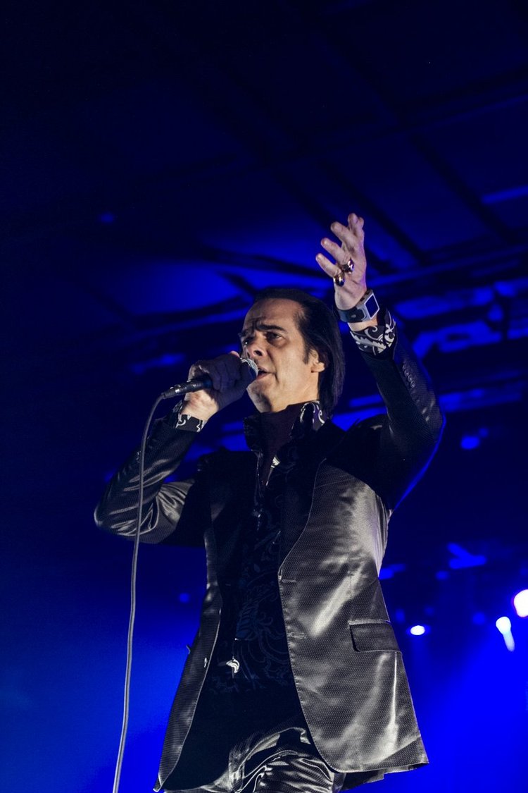 A male singer performing on stage under blue lighting, holding a microphone in his right hand and gesturing with his left hand.