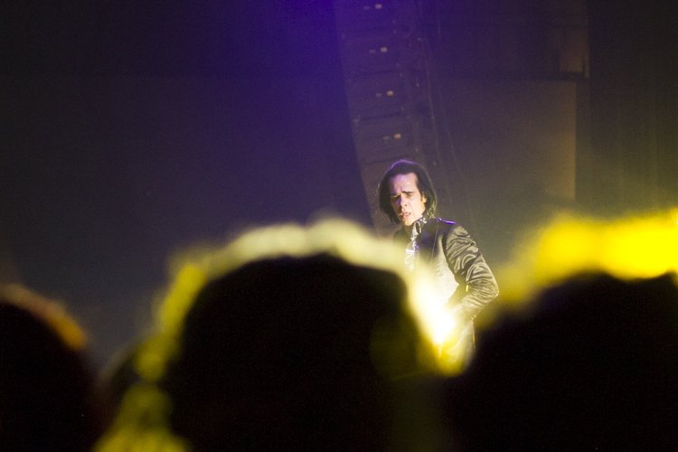 A performer with dark shoulder-length hair and a dark outfit is on stage under yellow and purple lighting, with an audience in front.
