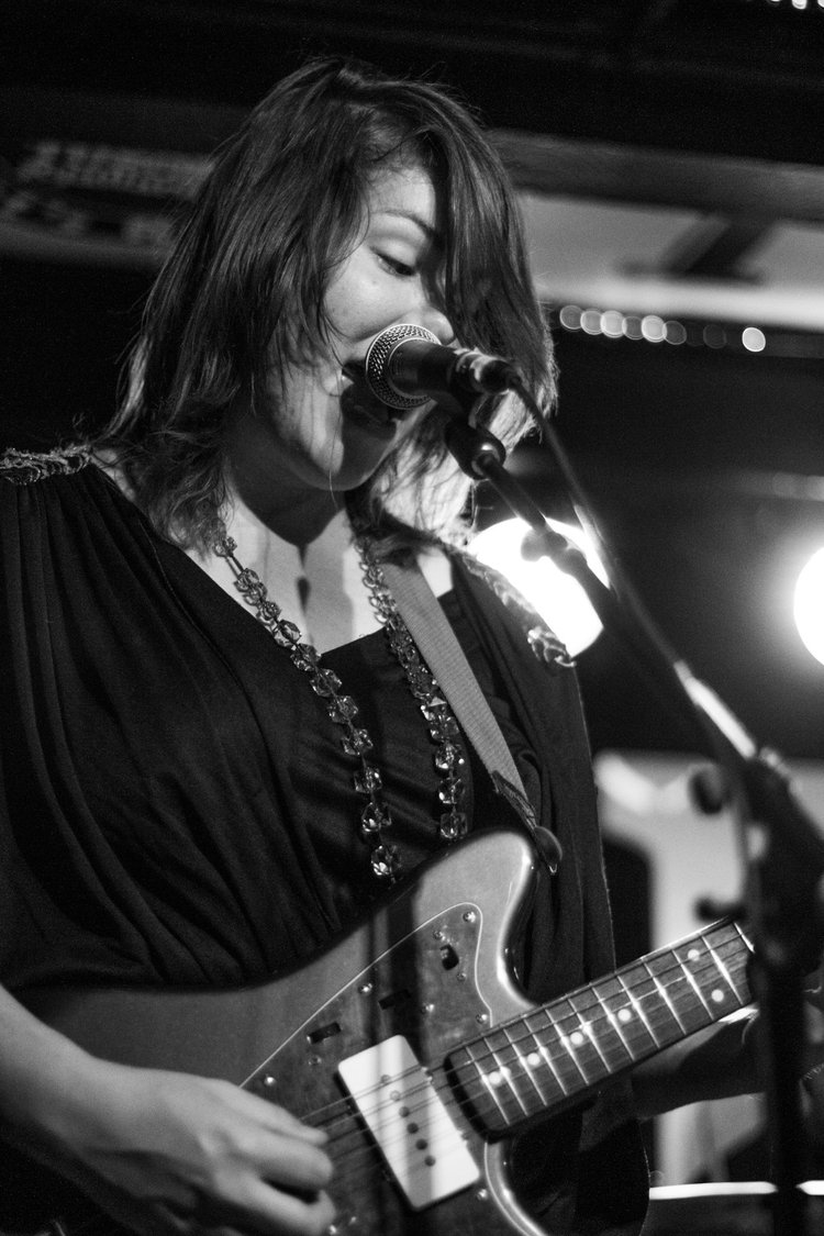 A woman singing into a microphone while playing an electric guitar at a live performance, in black and white.