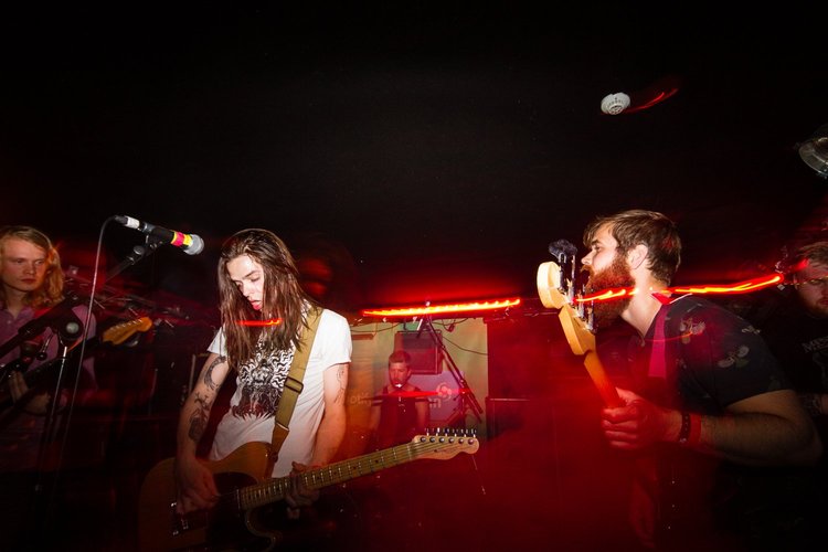 A band performing live in a dimly lit venue, with a woman playing guitar, a man singing with a guitar, and other band members in the background.